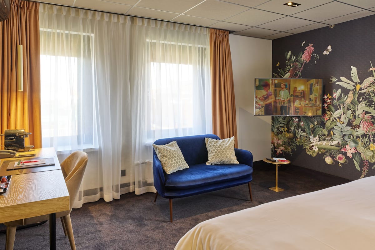 A hotel room with a bed, a blue sofa, a wooden desk, and a painting on the wall.