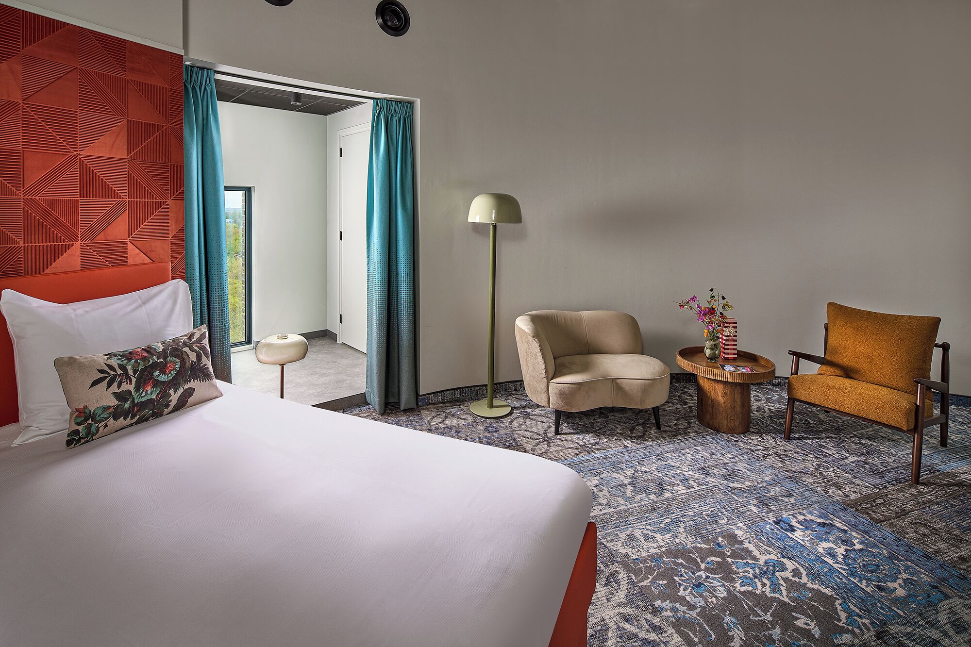Une chambre d’hôtel comprenant un lit, deux fauteuils, une lampe de sol, une table d’appoint avec un vase et un tapis à motifs.