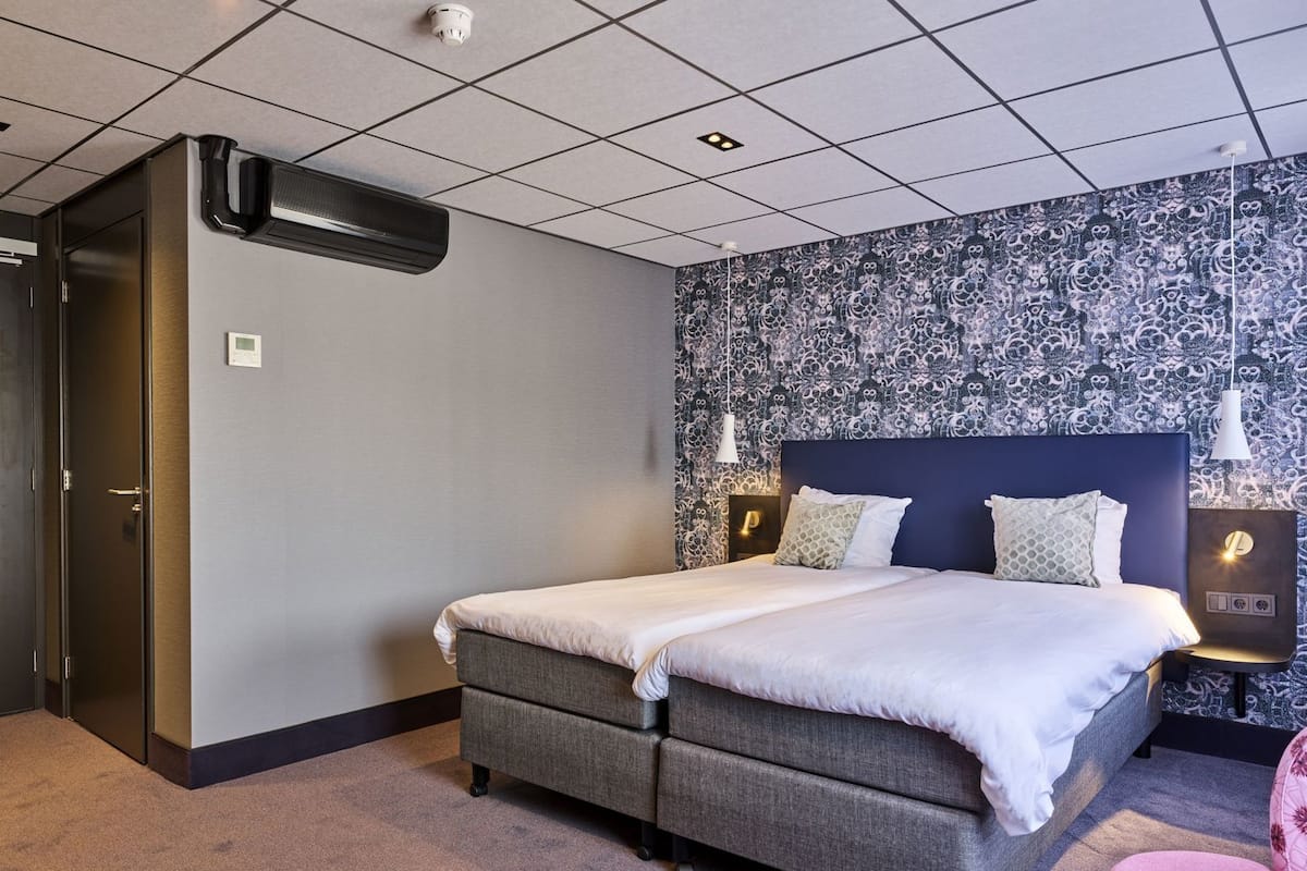 A hotel room with a bed, a wall-mounted air conditioner, a patterned wallpaper, and a small table with a lamp.