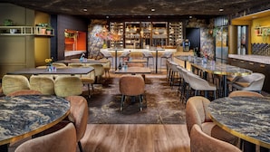 Un restaurante con mesas redondas y sillas acolchadas, una zona de bar y plantas decorativas.