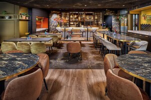 Un restaurante con mesas redondas y sillas acolchadas, una zona de bar y plantas decorativas.