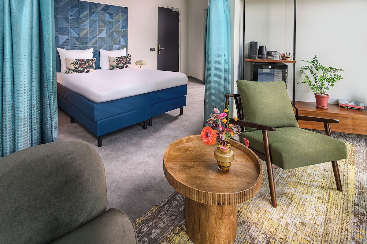 A hotel room with a bed, a chair, a small table with a vase of flowers, and a desk with a coffee machine.