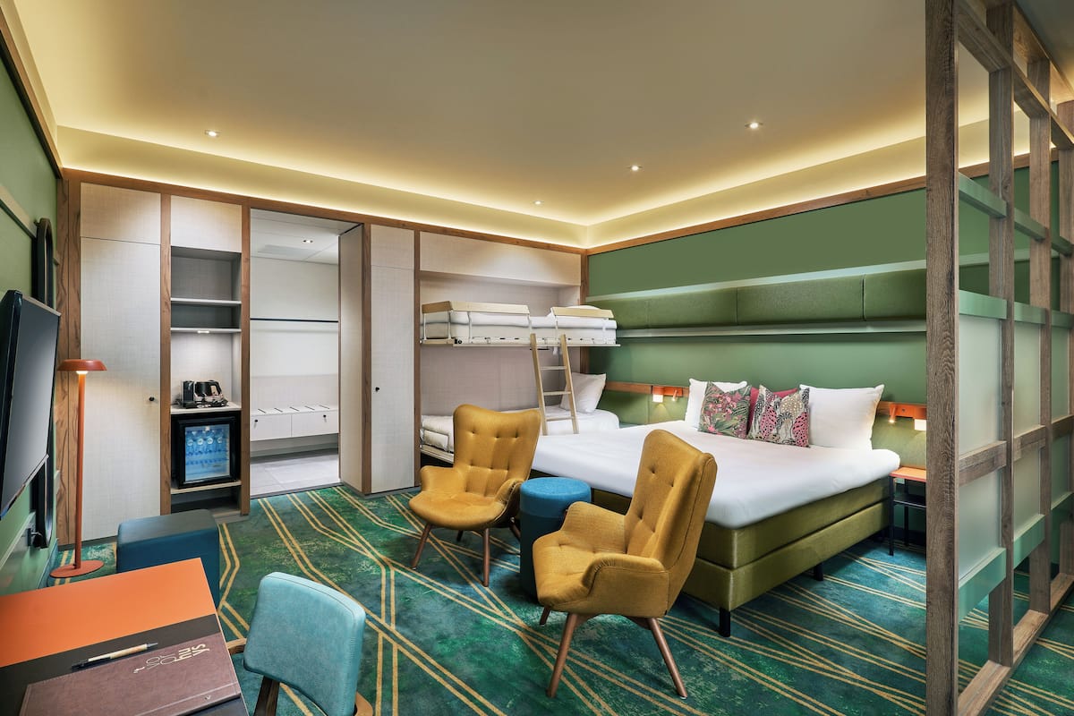 A hotel room with a bed, two armchairs, a desk, a wardrobe, and a television.