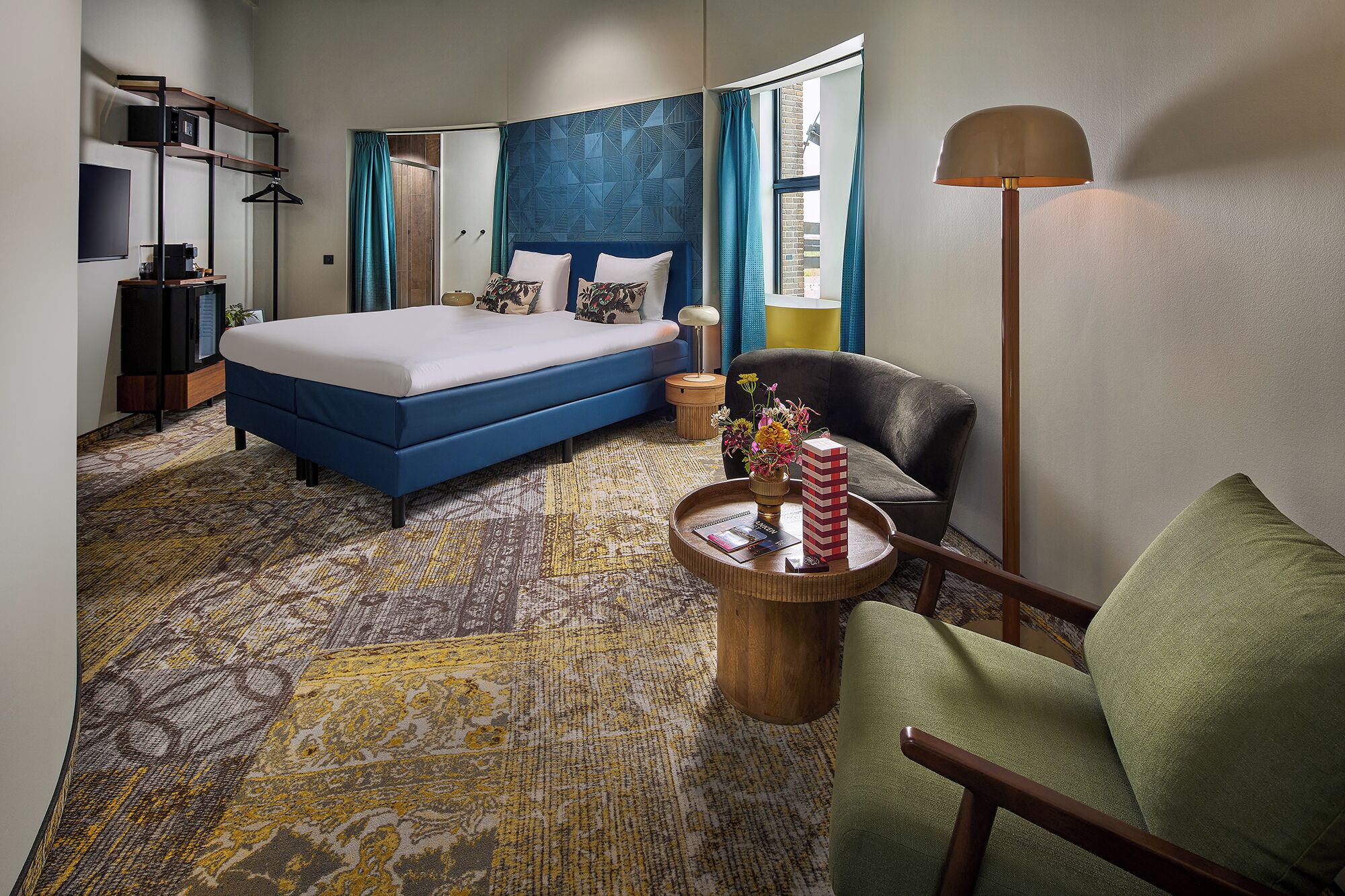 A hotel room with a bed, a chair, a side table, a lamp, and a view of the outside.