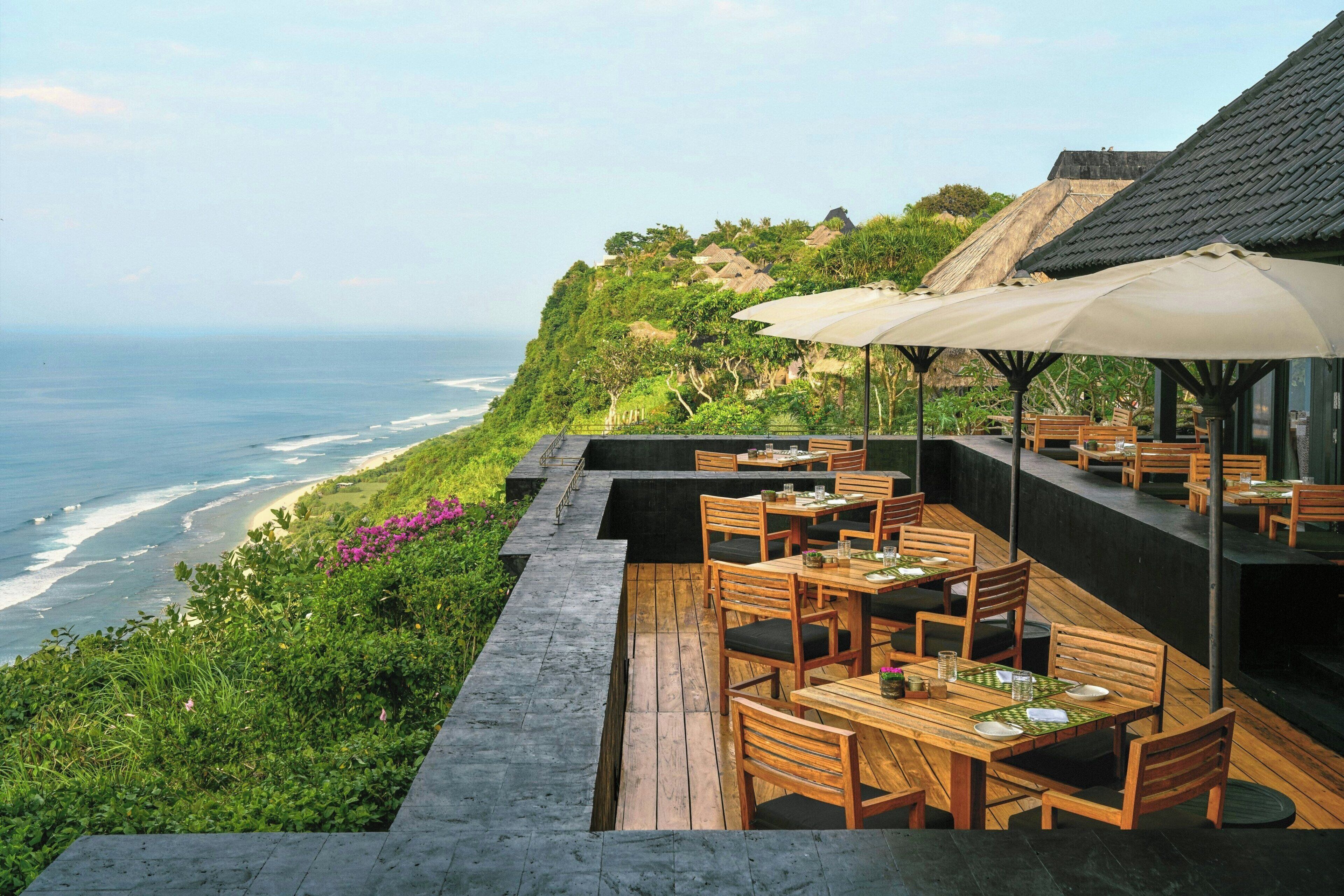 An outdoor dining area with wooden tables and chairs, overlooking a beach and ocean.