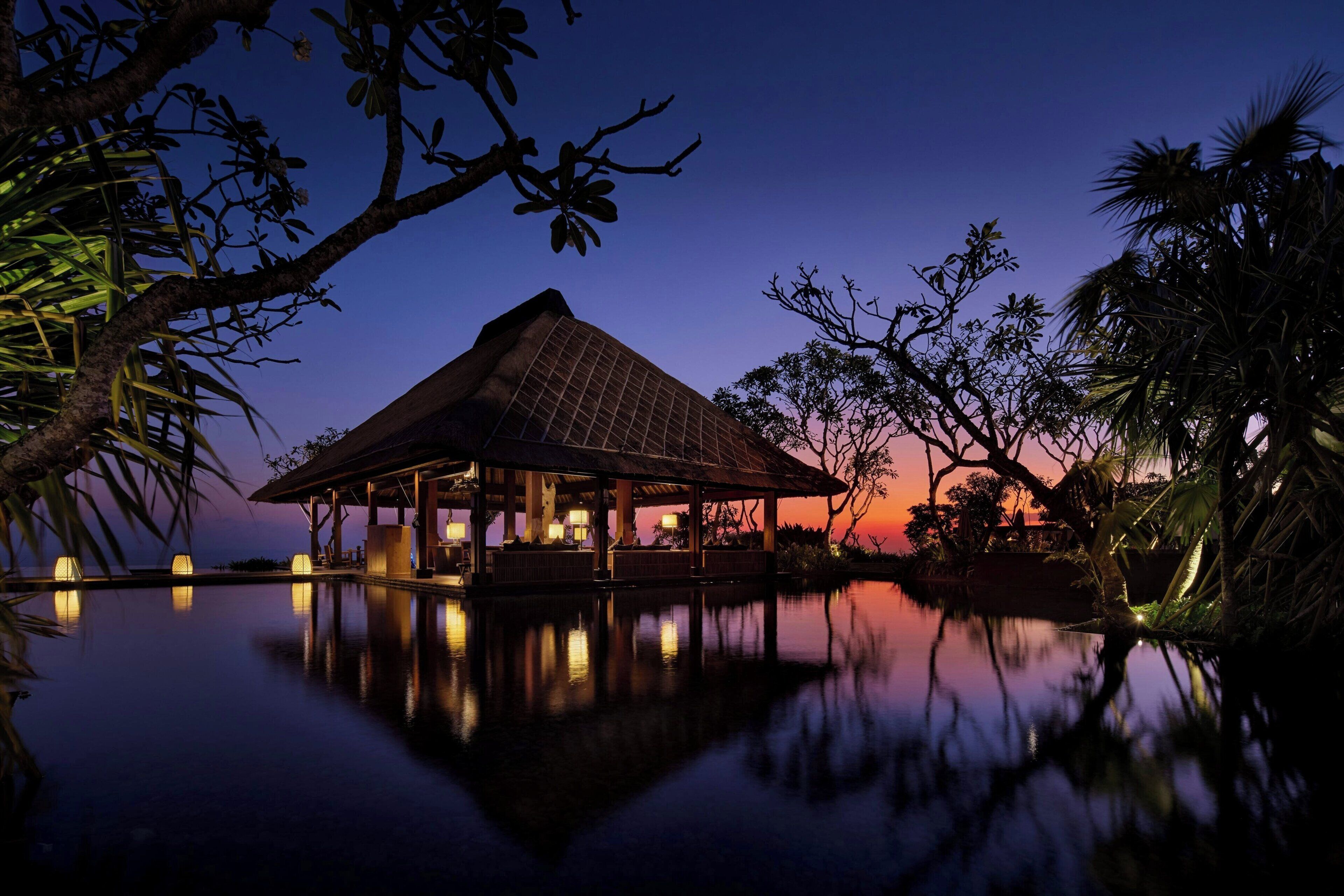 Escena tranquila al atardecer en un resort, con un pabellón de techo de paja sobre una piscina que refleja el entorno, rodeado de abundante vegetación tropical.