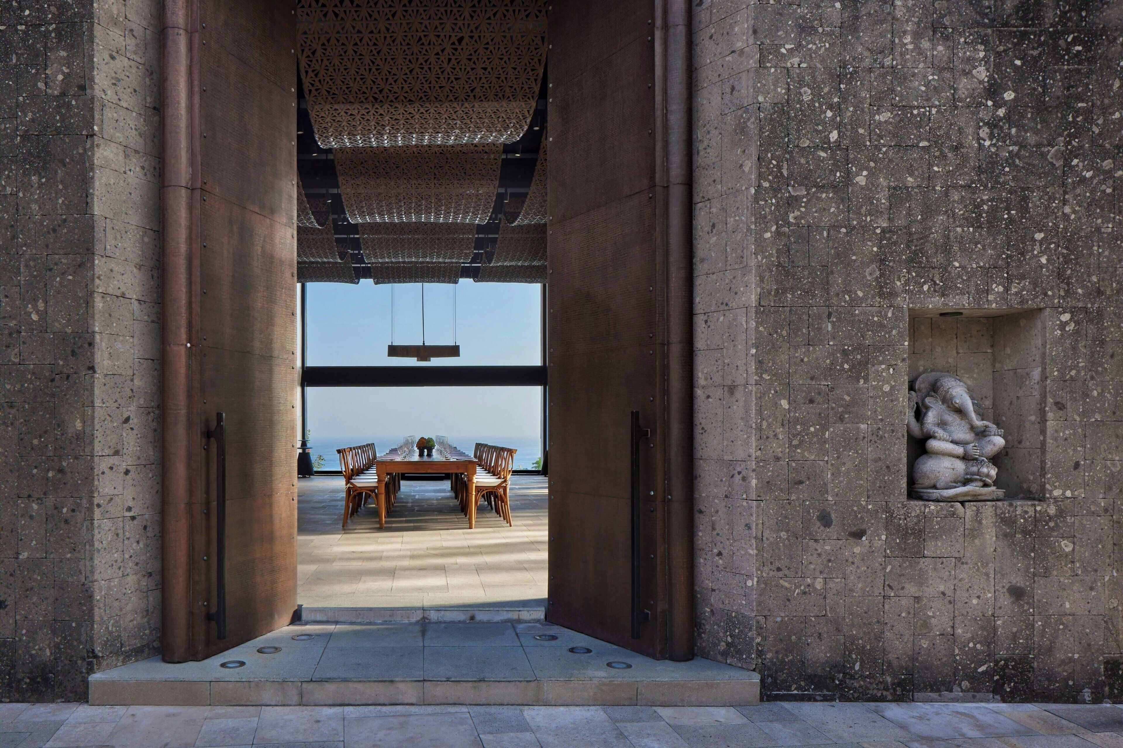 On aperçoit, à travers une grande porte ouverte, un coin repas avec une table et des chaises, un luminaire suspendu et une sculpture en pierre accrochée au mur.