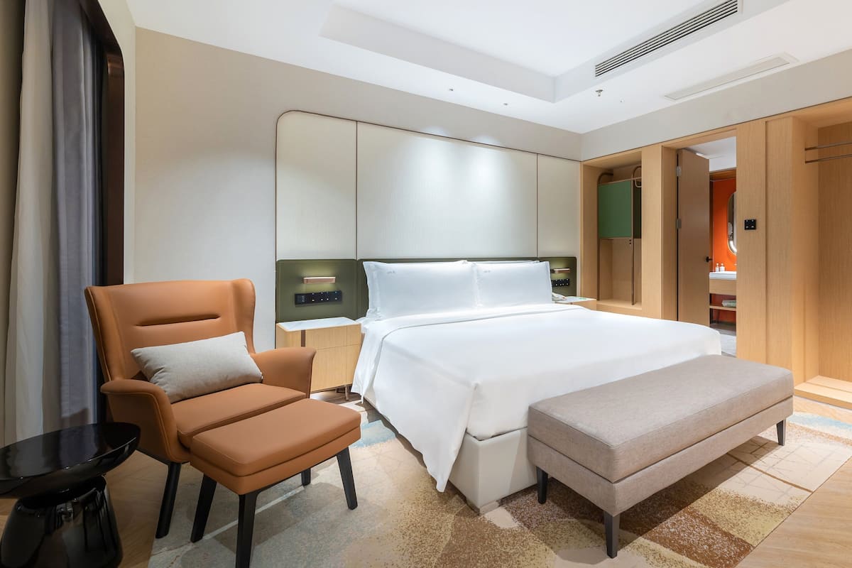 Een moderne hotelkamer met een groot bed, een zithoek met een stoel en een bankje, een nachtkastje en een kledingkast.