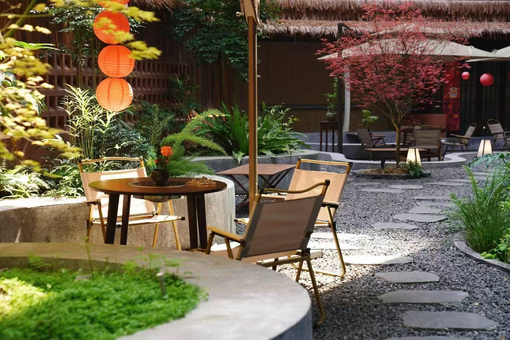 An outdoor seating area with tables and chairs, surrounded by plants and a thatched-roof structure.