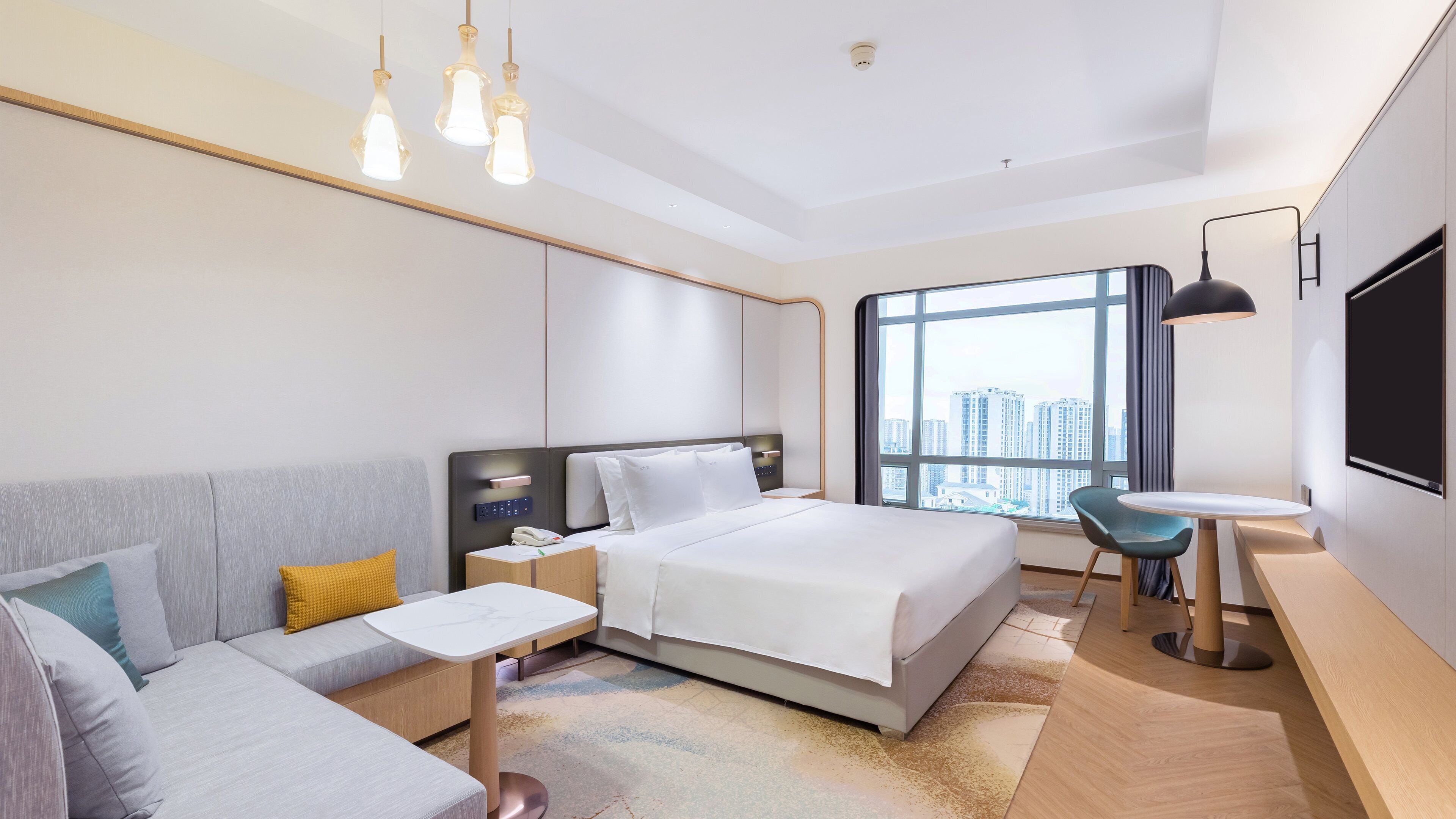A modern hotel room with a bed, a sofa, a desk, and a large window offering a city view.