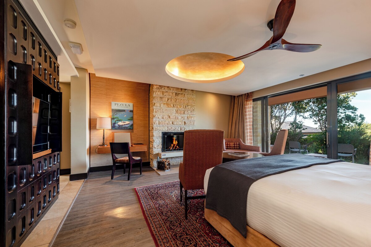 A hotel room with a large bed, a fireplace, a TV, and a balcony with a view of trees.