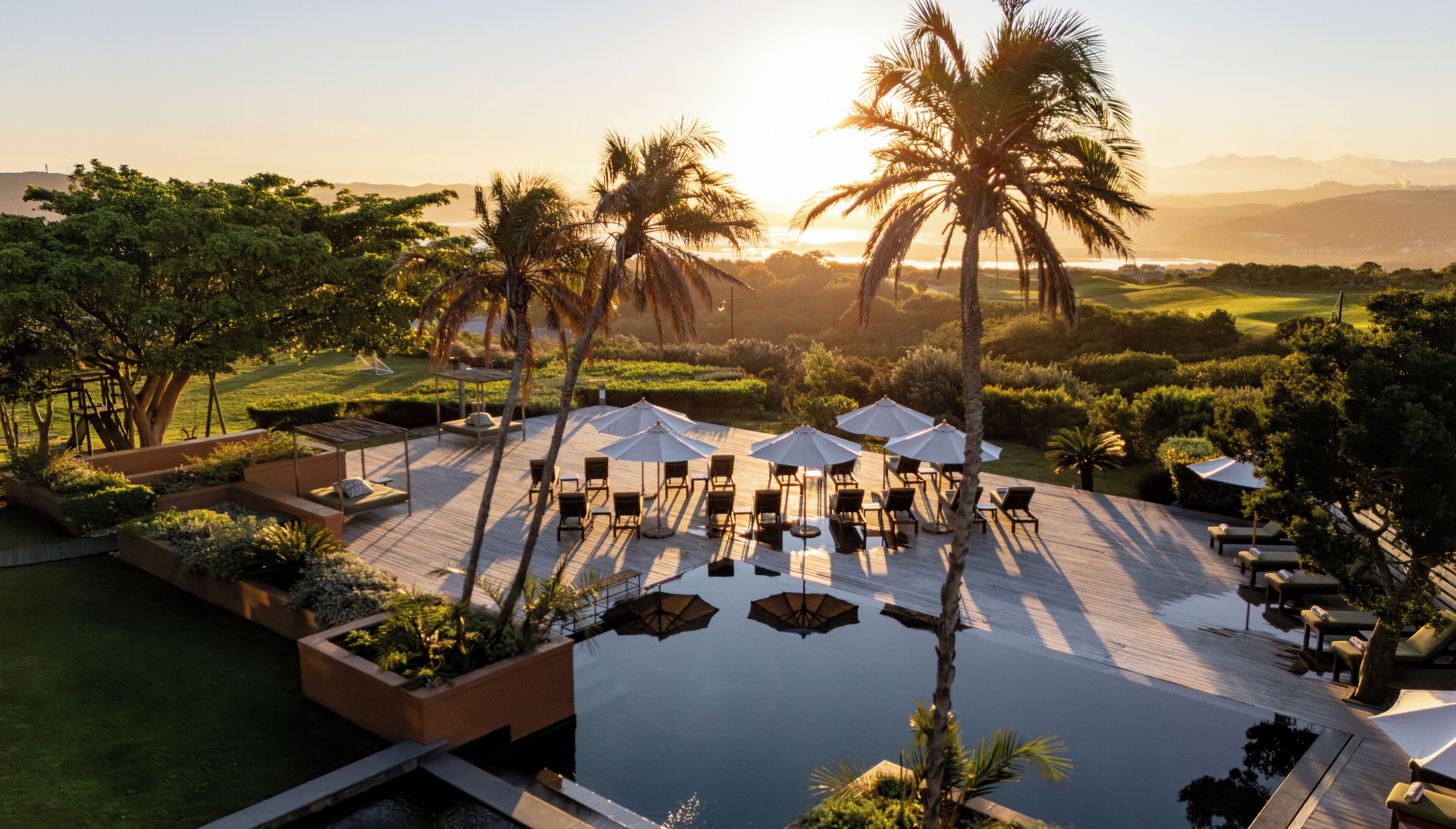 A resort pool area with lounge chairs and umbrellas, surrounded by palm trees and greenery.