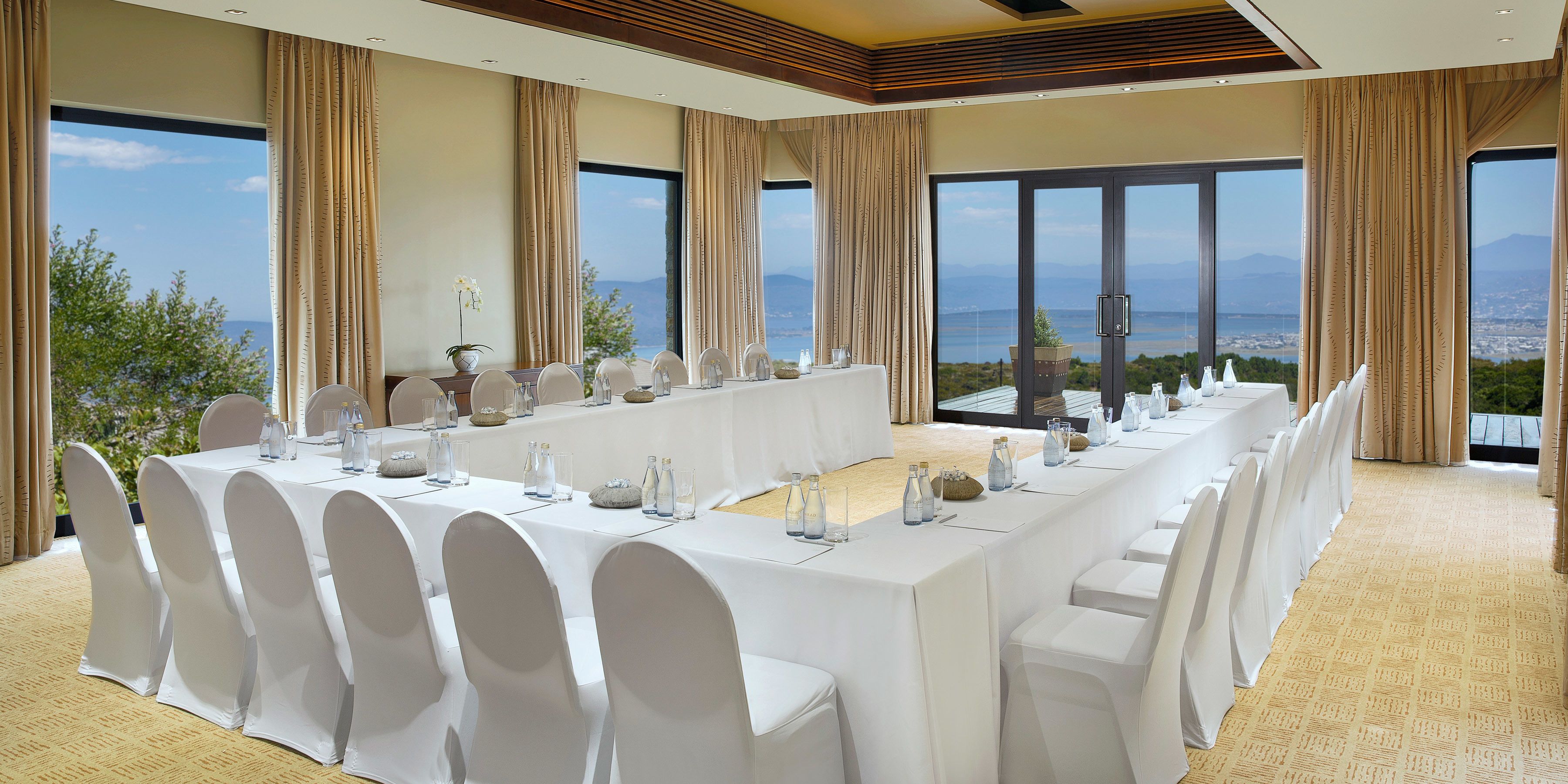 A conference room with a long table set for a meeting, large windows with a view of the sea and mountains, and a high ceiling with wooden paneling.