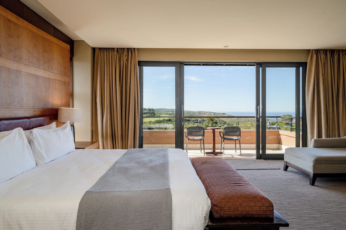 A hotel room with a large bed, a sofa, a balcony with a view of the countryside, and a dining area with chairs.