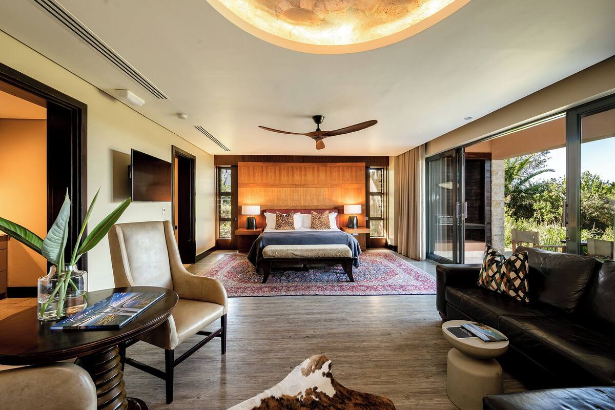 A modern hotel room with a large bed, a ceiling fan, a seating area with a sofa and armchairs, and a view through a glass door.