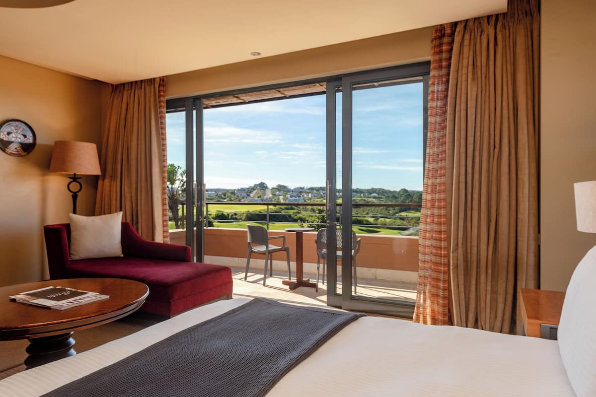 A hotel room with a large bed, a red armchair, a small table, and a balcony with a view of the countryside.