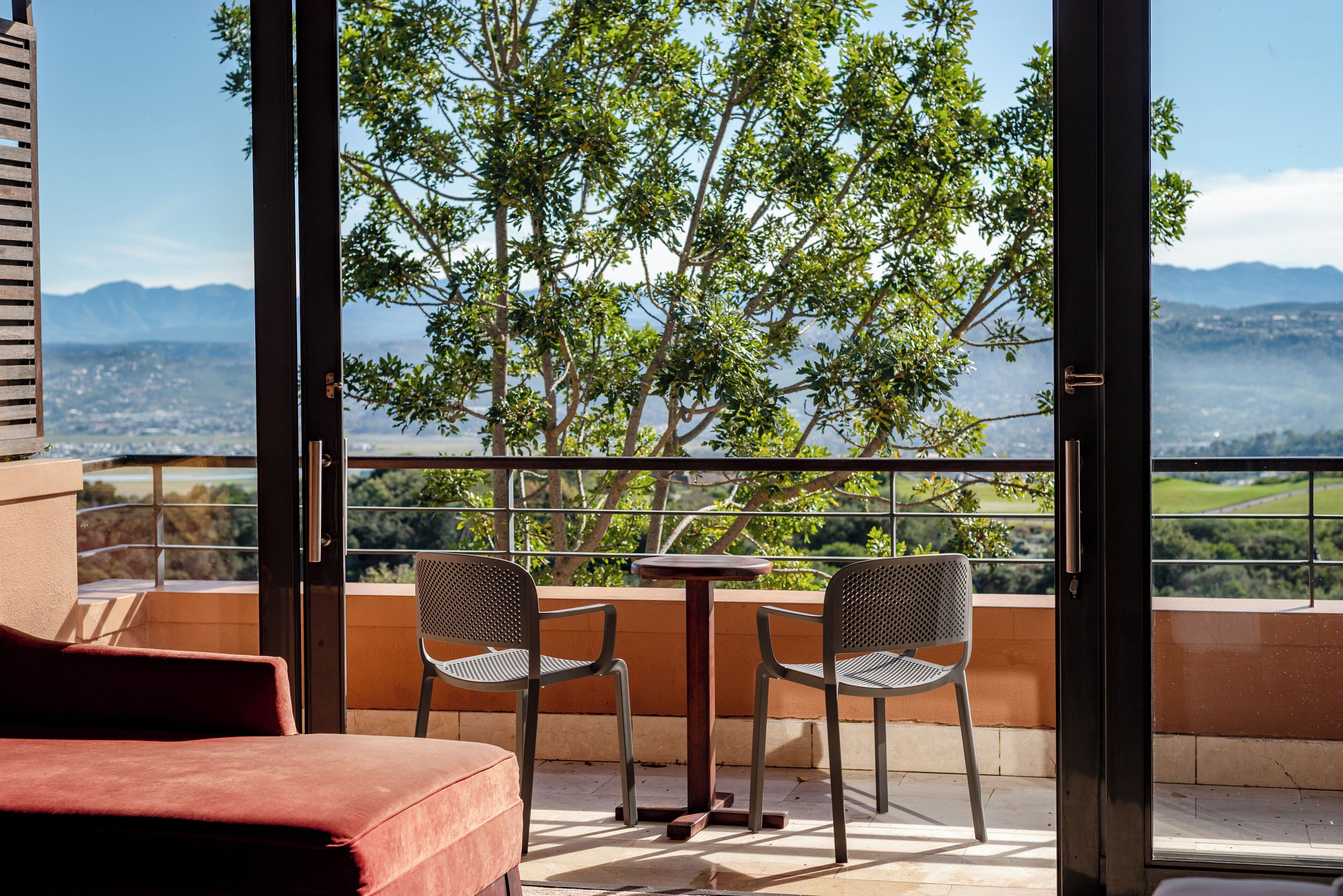 A balcony with a table and two chairs, overlooking a scenic view of trees and distant mountains.