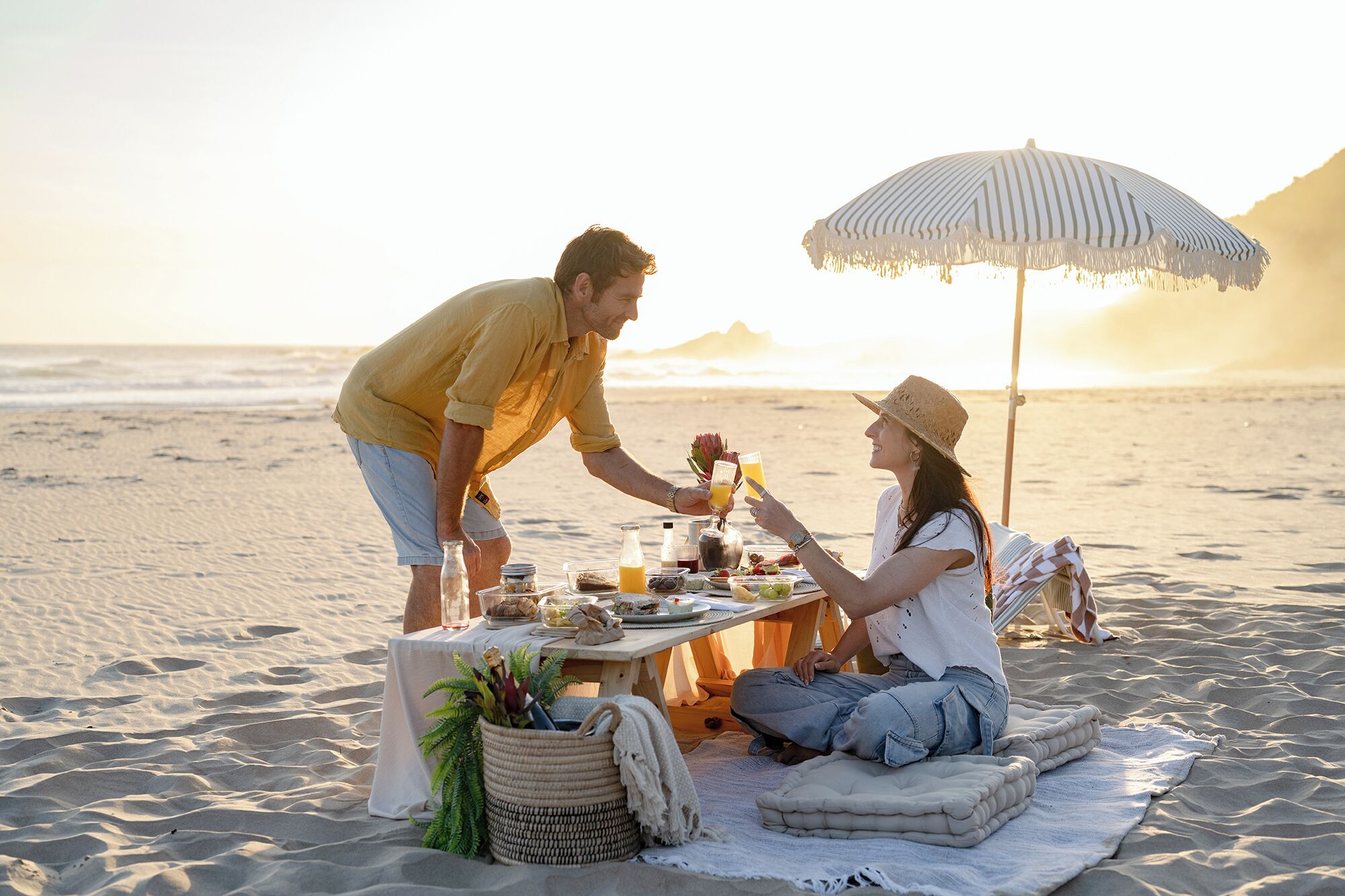 Un couple pique-nique sur une plage de sable, une table est dressée pour deux, une couverture est étalée, et un grand parasol offre de l’ombre.