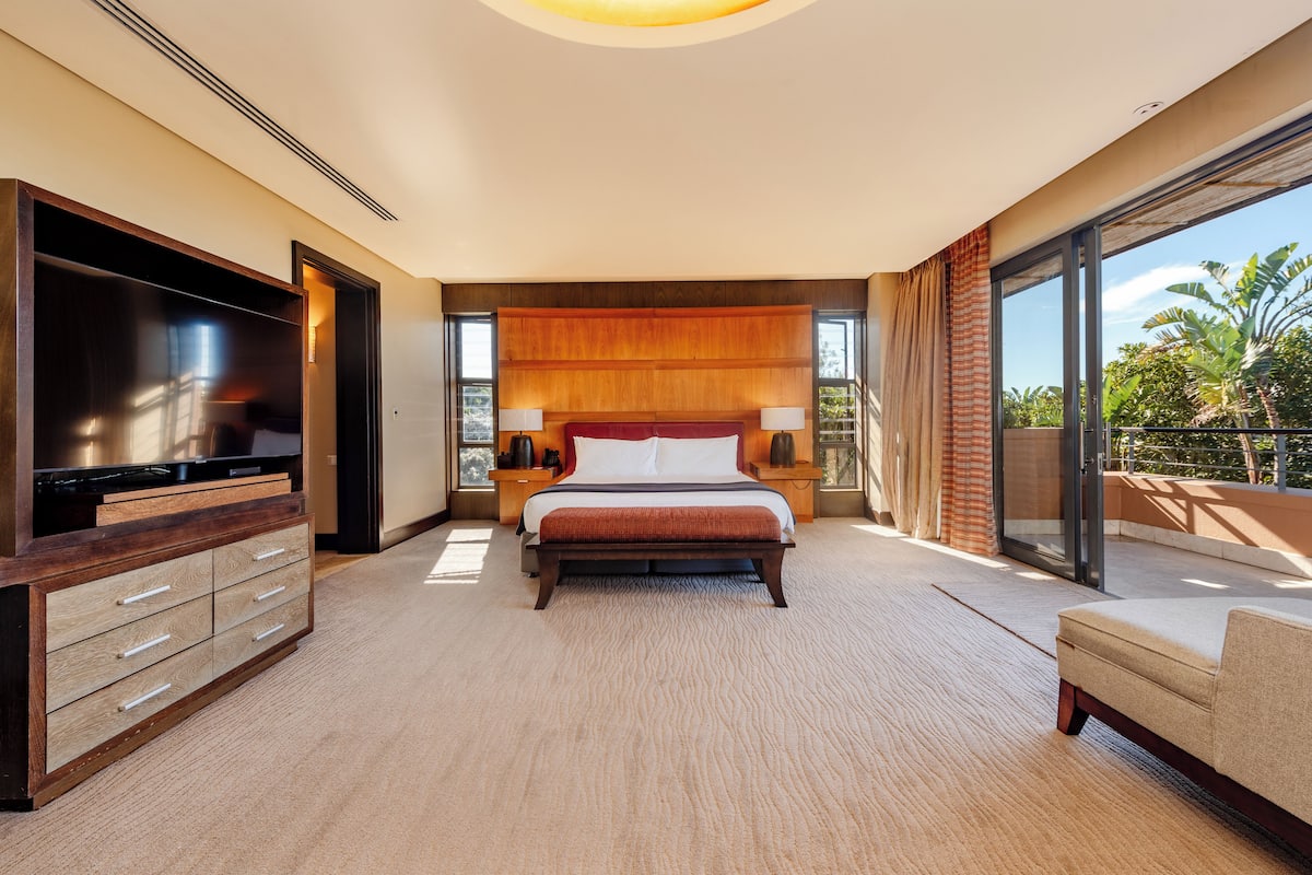 A spacious hotel bedroom with a large bed, a flat-screen TV, a wooden dresser, and a balcony with a view of palm trees.