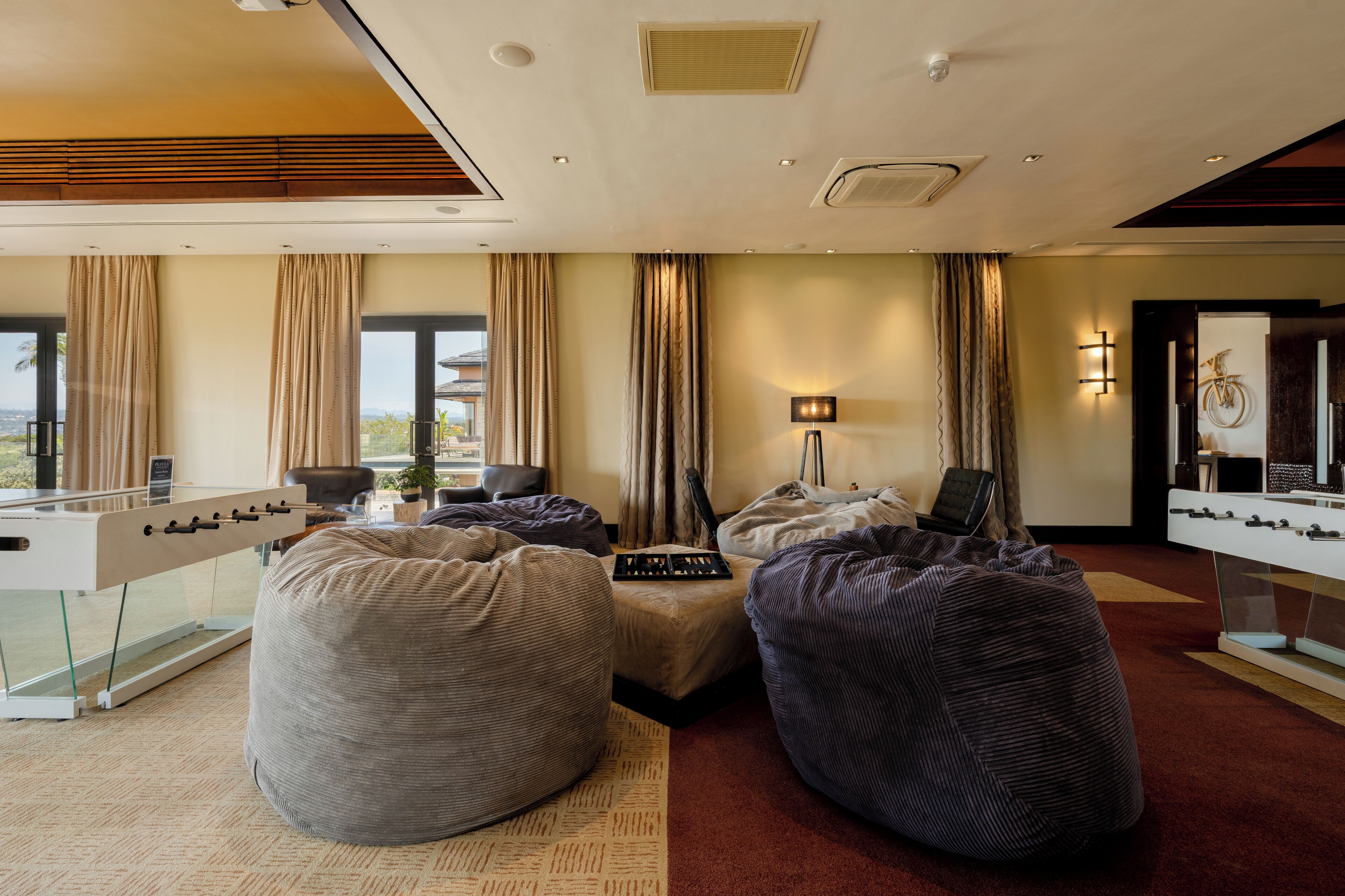 A modern hotel room with a foosball table, bean bag chairs, a desk with a laptop, and a large window offering a view.