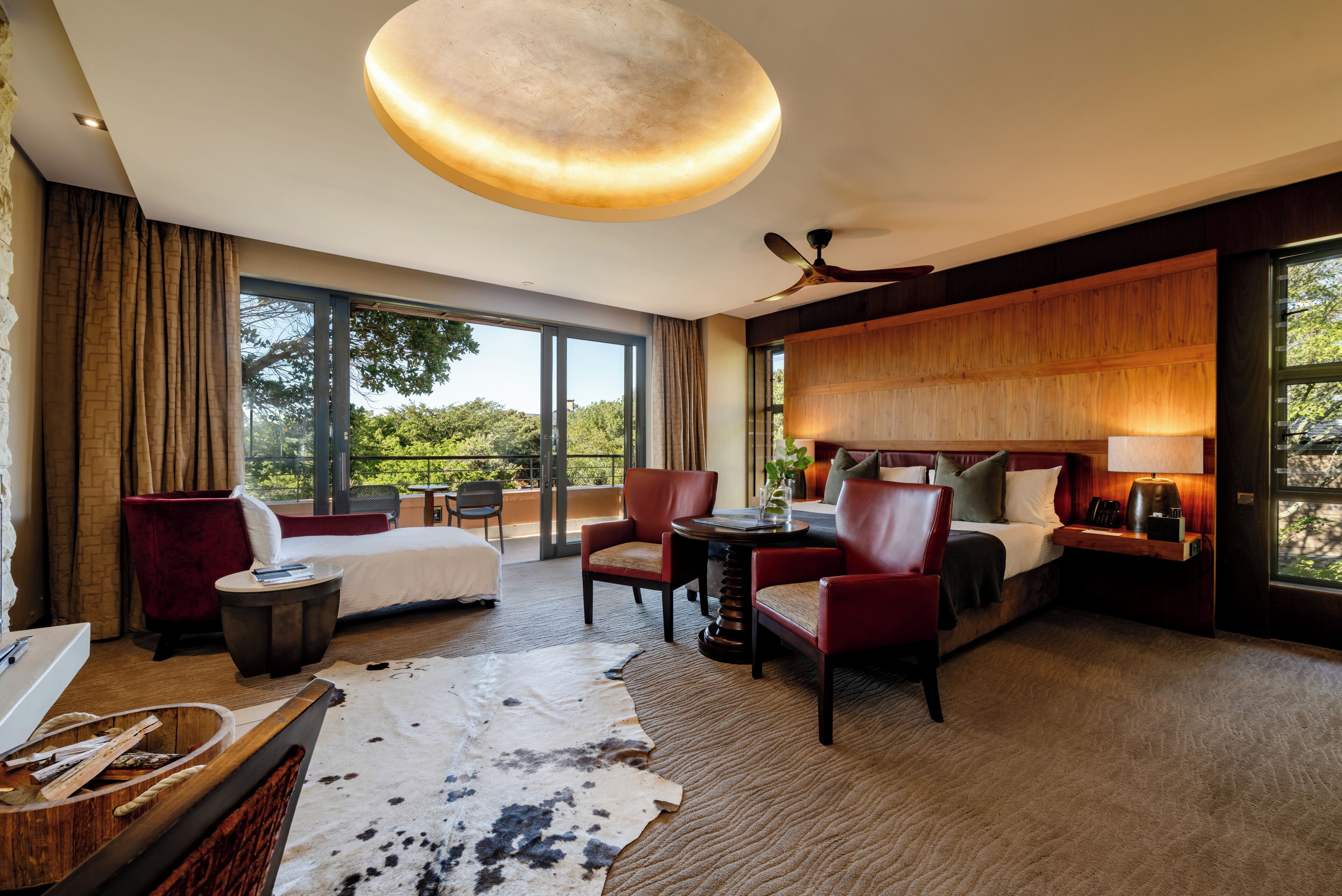 Amplia habitación de hotel con una cama grande, una zona de estar con sillas, una mesa de comedor, un ventilador de techo y grandes ventanales con vistas al exterior.