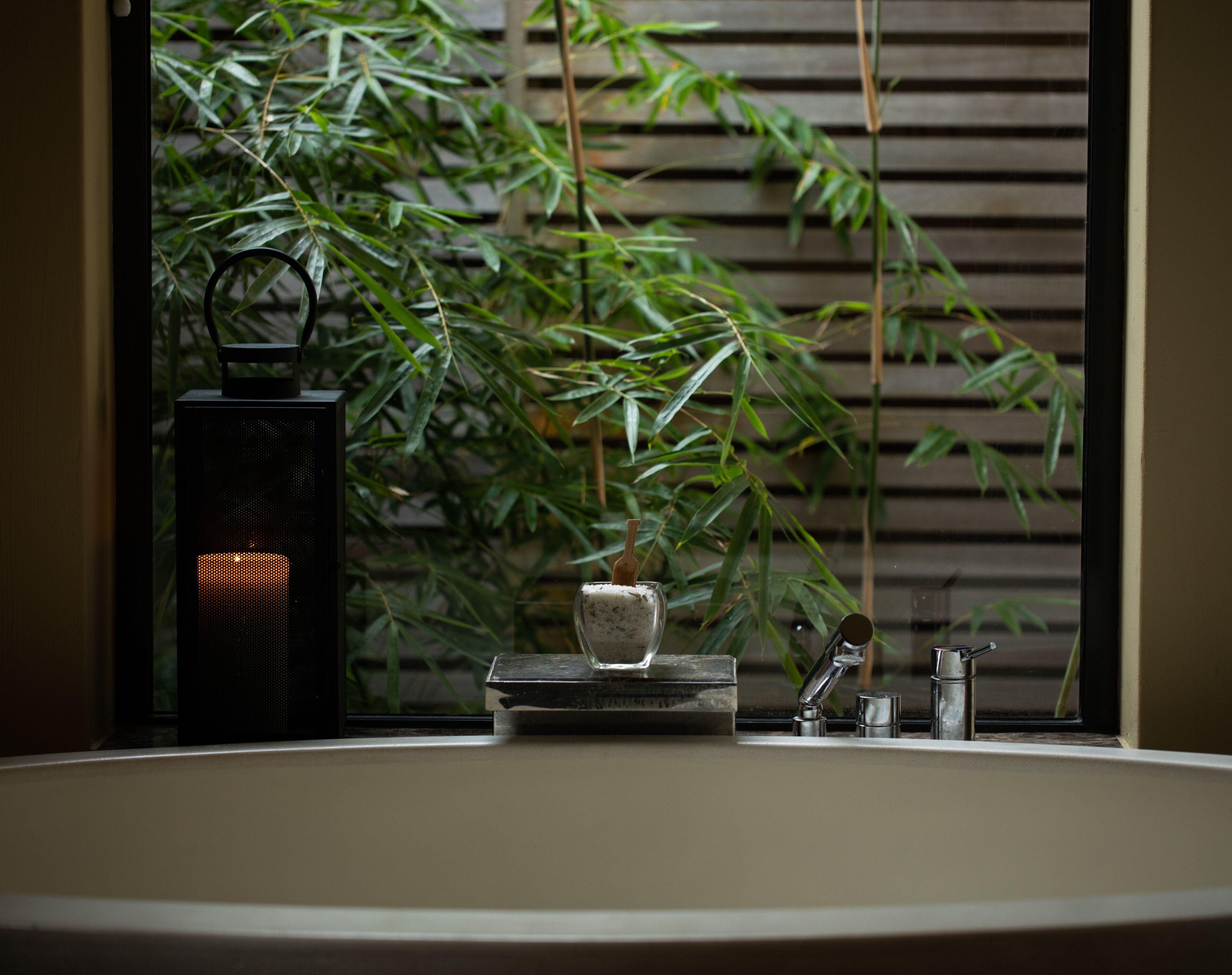 A bathroom with a sink, a small table with a candle, and a window with bamboo plants.