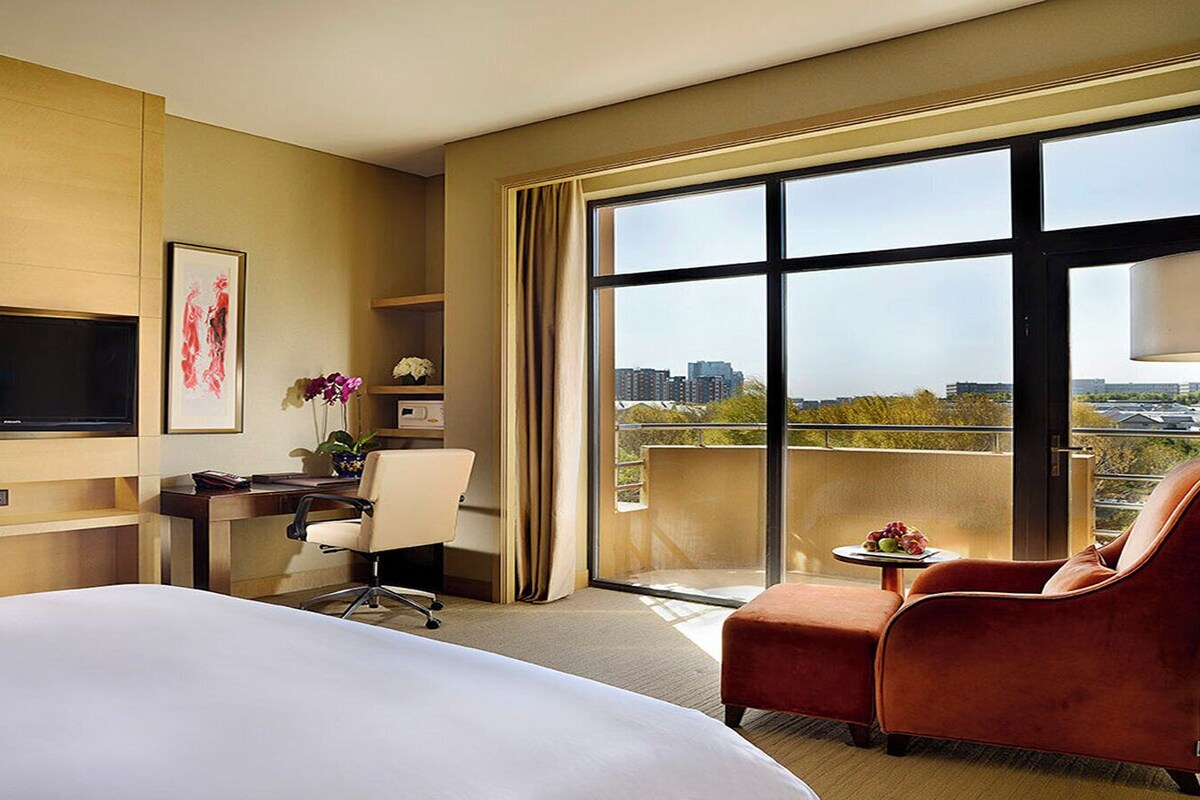 A hotel room with a large bed, a desk with a chair, a TV mounted on the wall, and a balcony with a view of the city.
