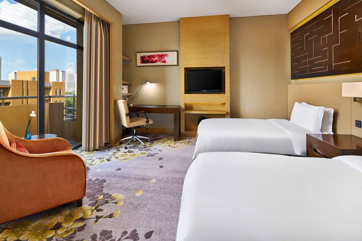 A hotel room with two beds, a desk, a chair, a TV, and a large window with a city view.