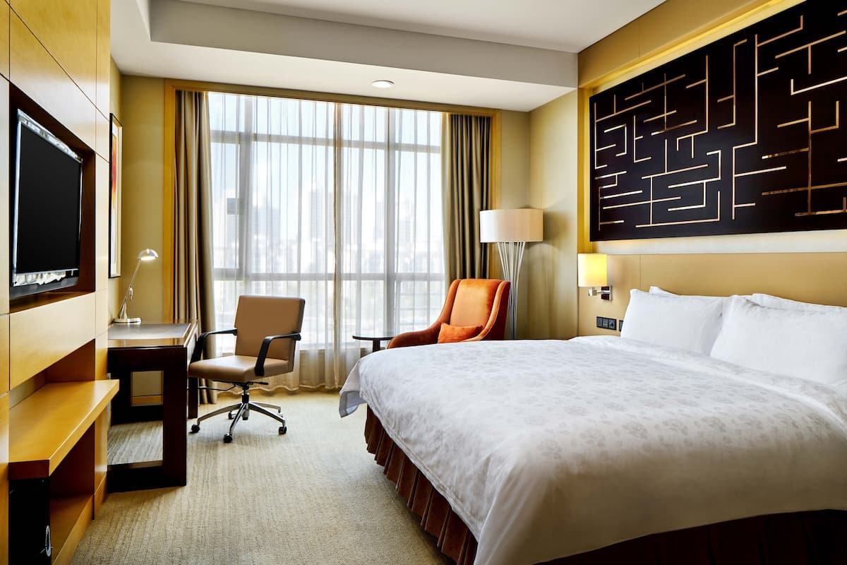 A hotel room with a large bed, a desk with a chair, a television, and a large window with curtains.