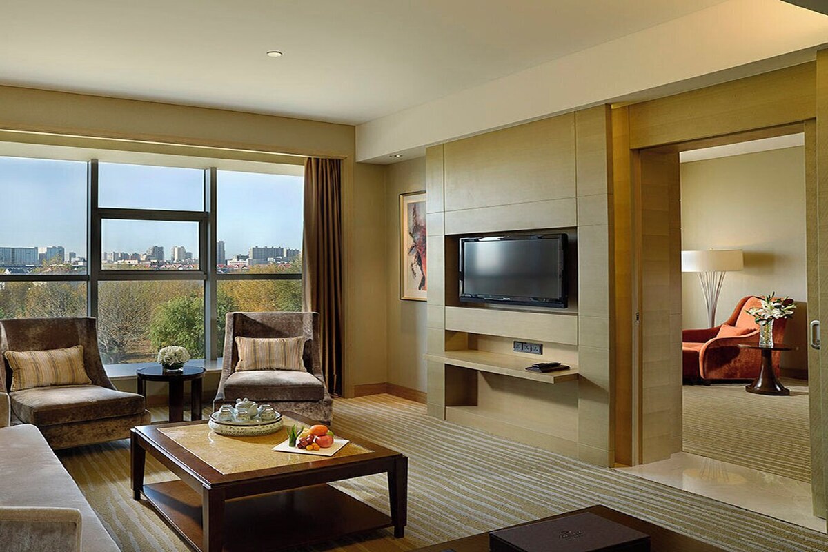 A modern hotel room with a sofa, two armchairs, a coffee table with fruit, a TV, and a city view.