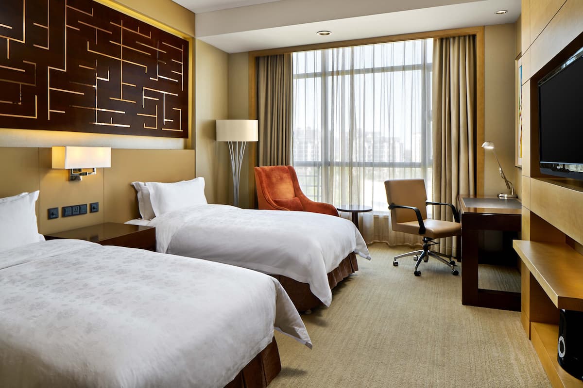 A hotel room with two beds, a desk, a TV, and a large window with curtains.