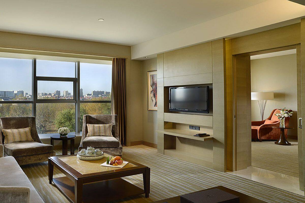 A spacious hotel room with a large window offering a city view, a flat-screen TV, a sofa, two armchairs, and a coffee table with a decorative tray.