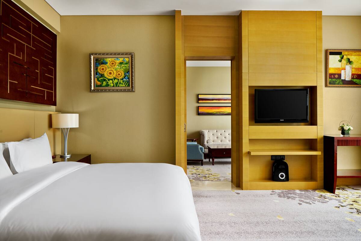 A hotel room with a large bed, a television, a painting, and a sofa in the background.
