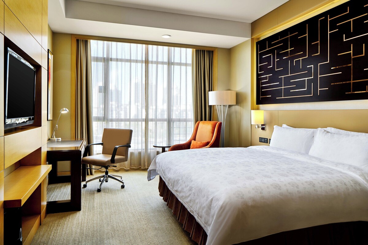 A hotel room with a bed, desk, chair, TV, and a large window with curtains.