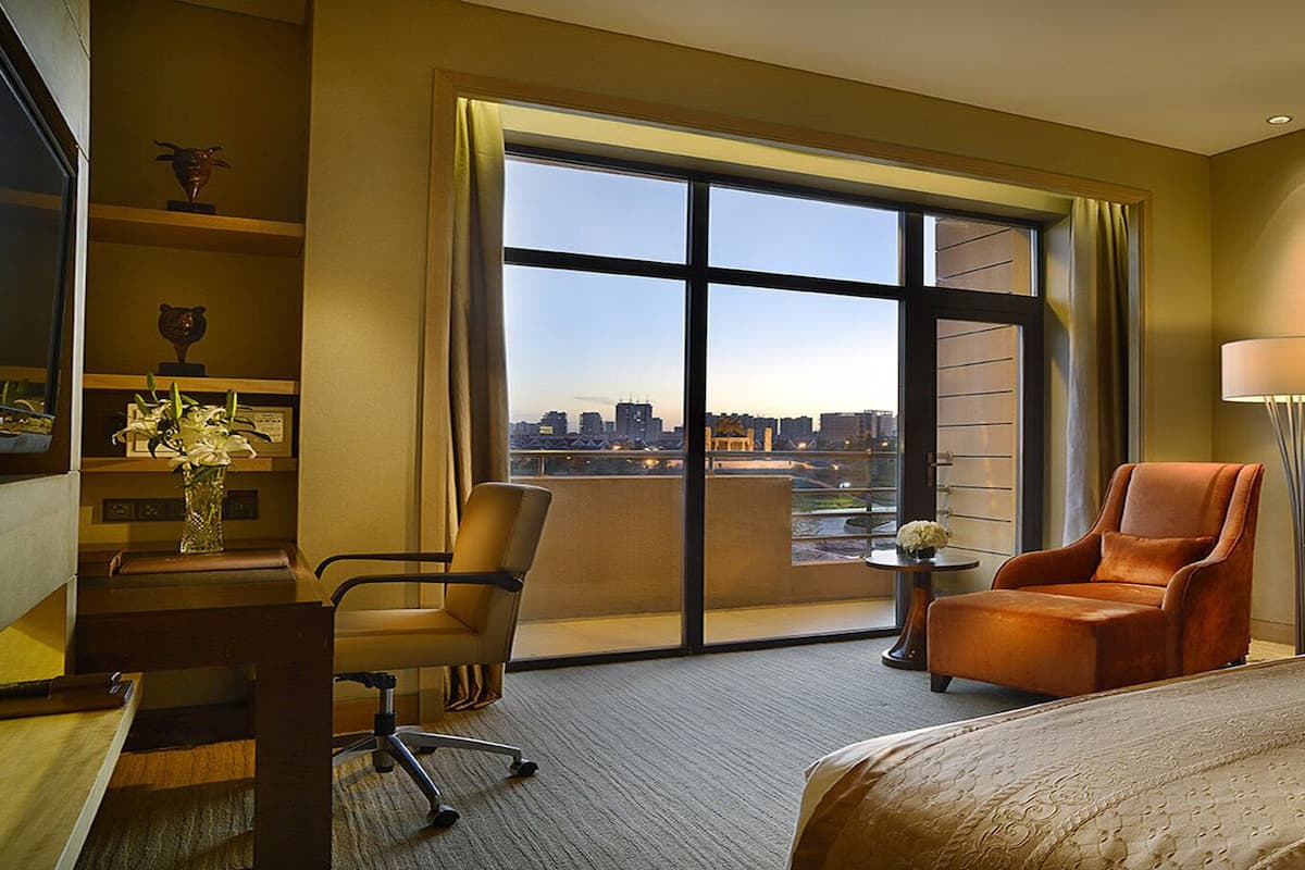 A hotel room with a large window, a bed, a desk, a chair, and a lamp.