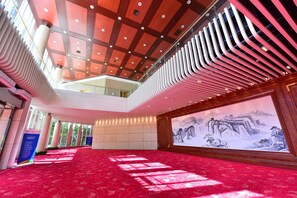 A spacious hall with a red carpet, a high ceiling with modern lighting, and a large wall-mounted painting of a mountainous landscape.