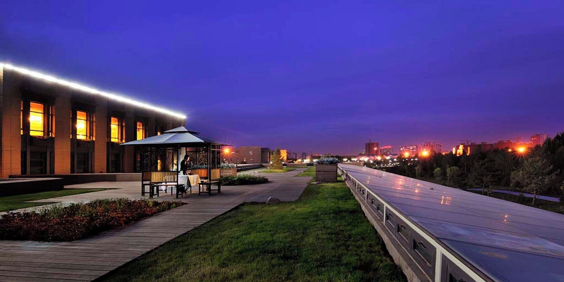 A covered outdoor seating area with tables and chairs, a building with illuminated windows, and a pathway leading to a bridge.