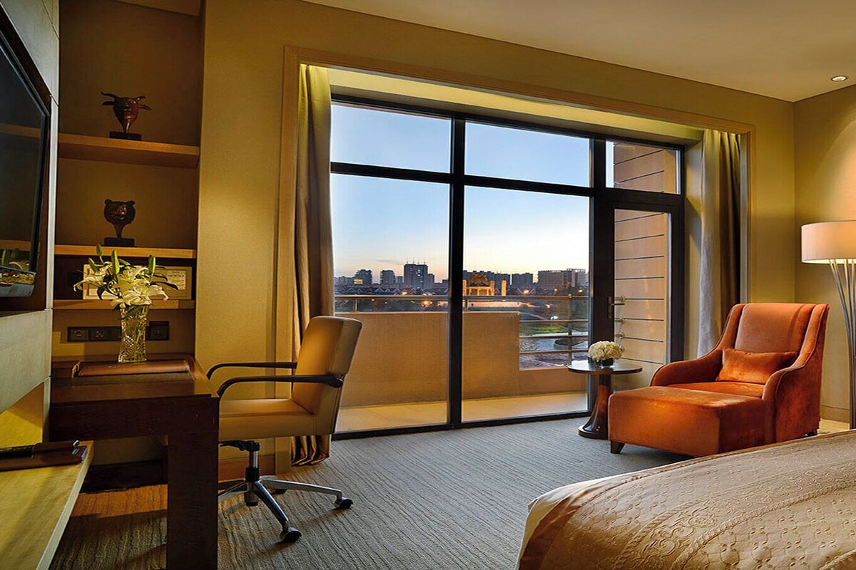A hotel room with a large window, a bed, a desk, a chair, and a lamp.