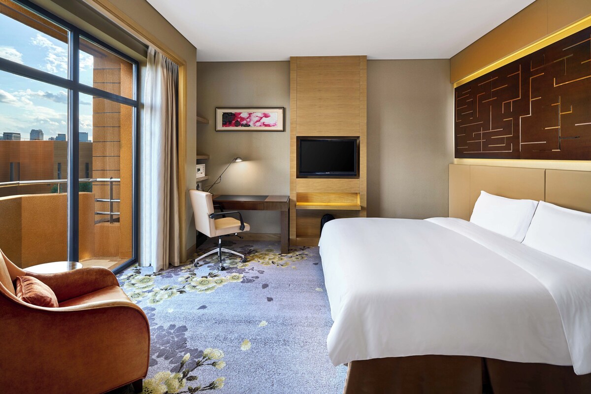 A hotel room with a bed, a desk, a chair, a TV, and a large window.