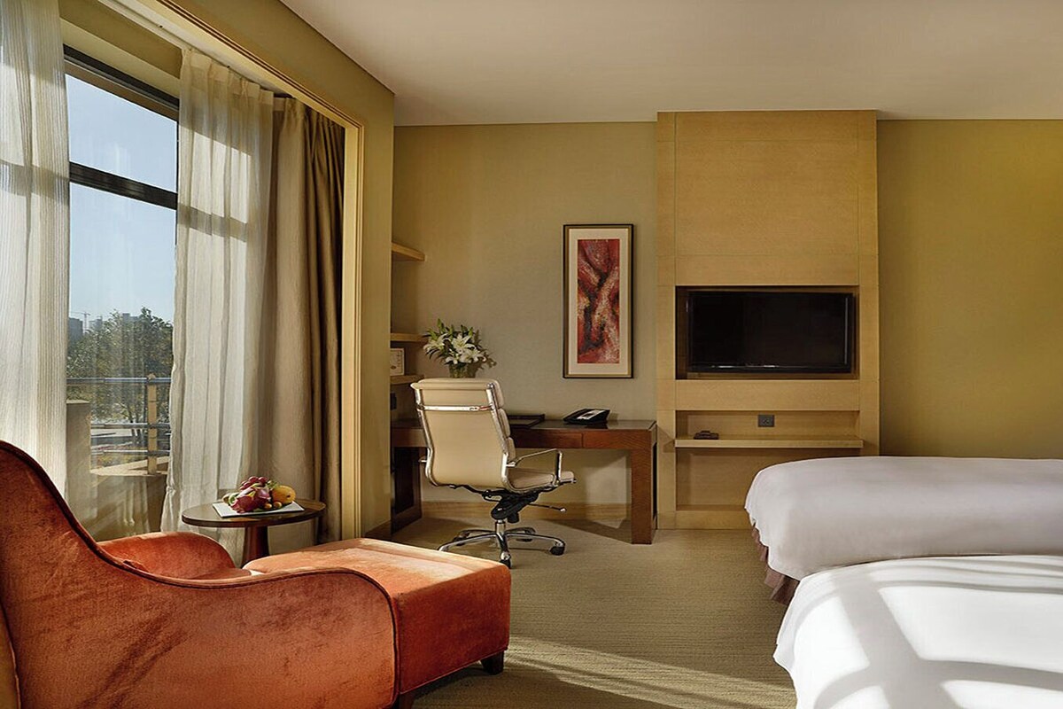 A hotel room with two beds, a desk, a TV, and a view of the outdoors.