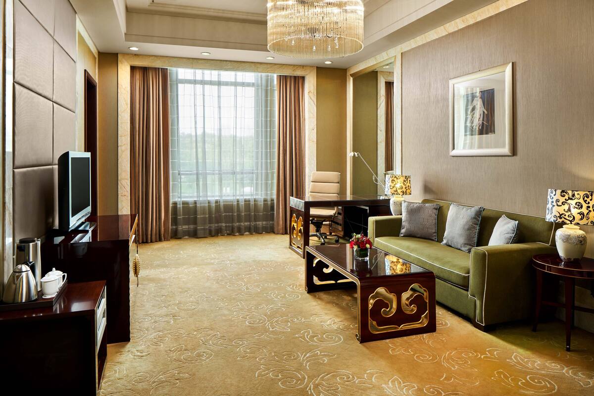 A modern hotel room with a large window, a green sofa, a wooden coffee table, and a television.