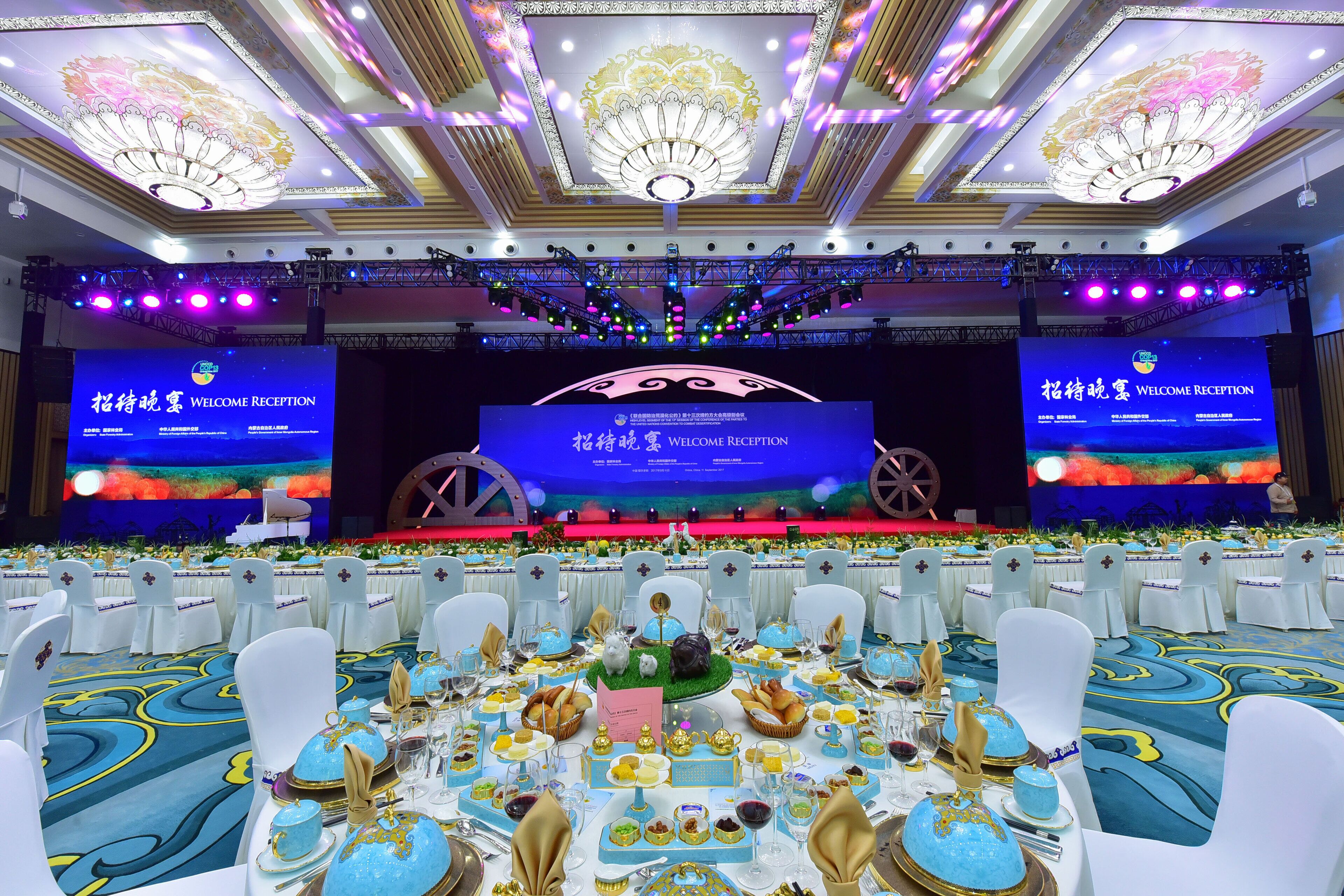 Un salón de banquetes con escenario, mesas dispuestas para una recepción y una pantalla grande que muestra el texto "WELCOME RECEPTION".