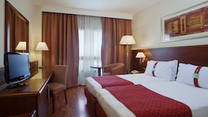 A hotel room with a bed, two bedside lamps, a desk with a television, a chair, and a window with curtains. - Holiday Inn Cagliari by IHG (Cagliari)
