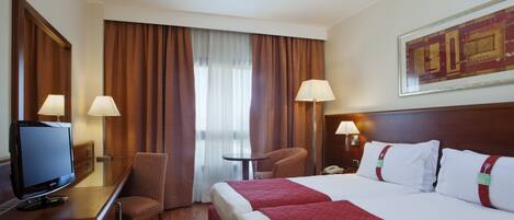 A hotel room with a bed, two bedside lamps, a desk with a television, a chair, and a window with curtains.