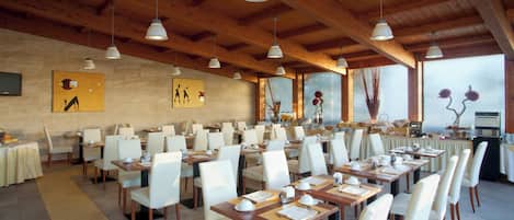 Un comedor amplio con altos techos de madera e iluminación colgante. Las mesas están dispuestas con manteles blancos y sillas, y hay obras de arte en las paredes.