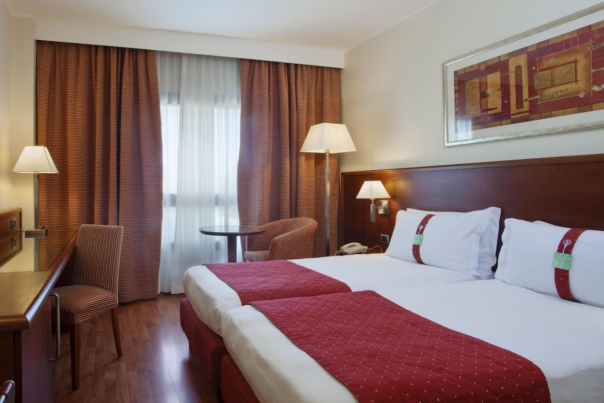 A hotel room with a large bed, two bedside tables with lamps, a desk with a chair, and a window with curtains.