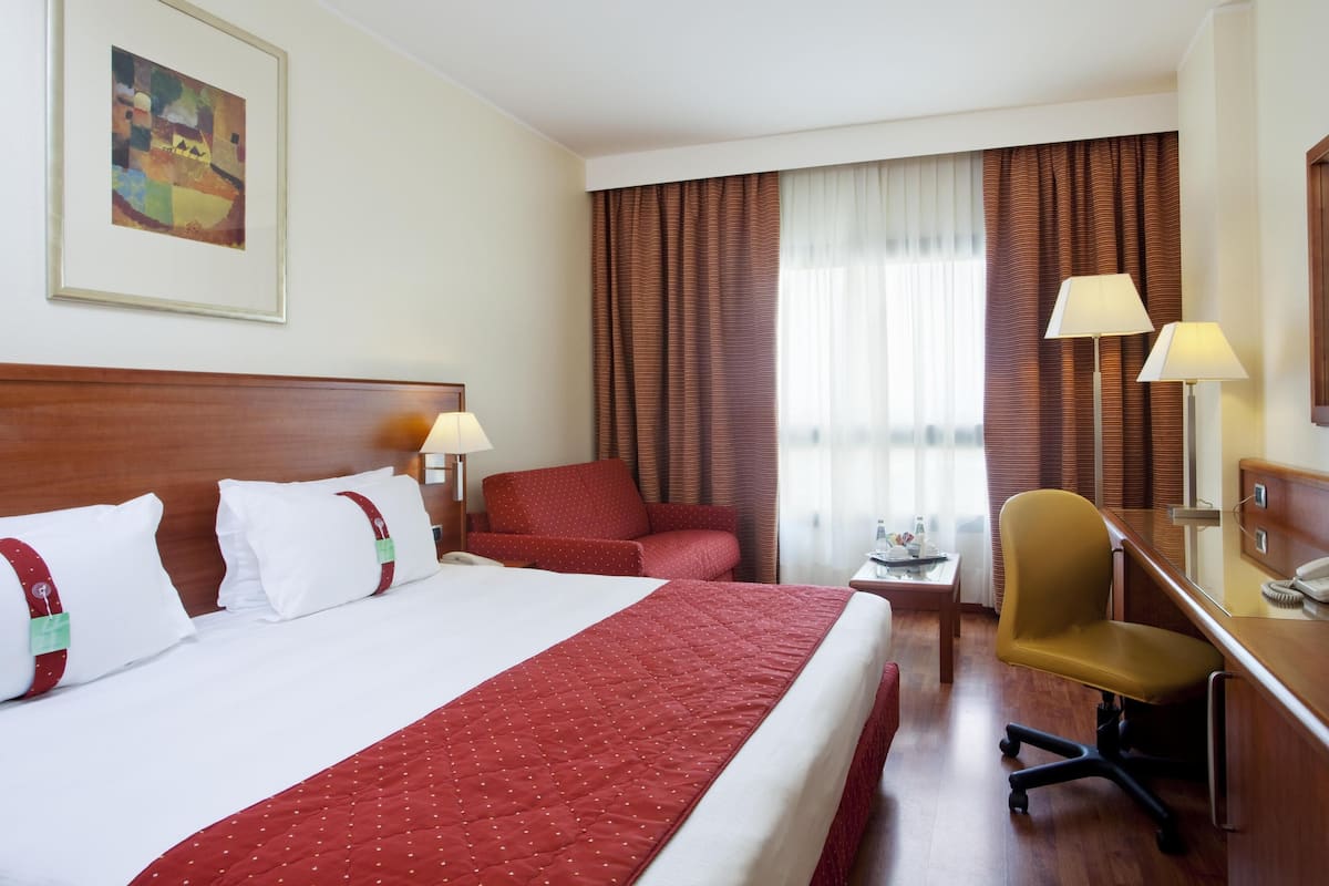 A hotel room with a large bed, a desk with a chair, a small table with a lamp, and a large window with curtains.