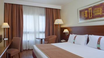 A hotel room with a large bed, a desk, a chair, a small table, two lamps, and a window with curtains.