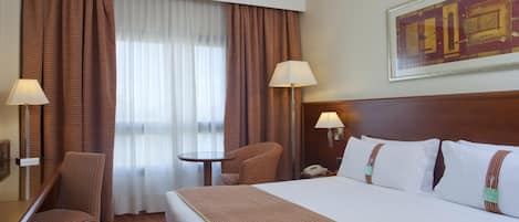 A hotel room with a large bed, a desk, a chair, a small table, two lamps, and a window with curtains.