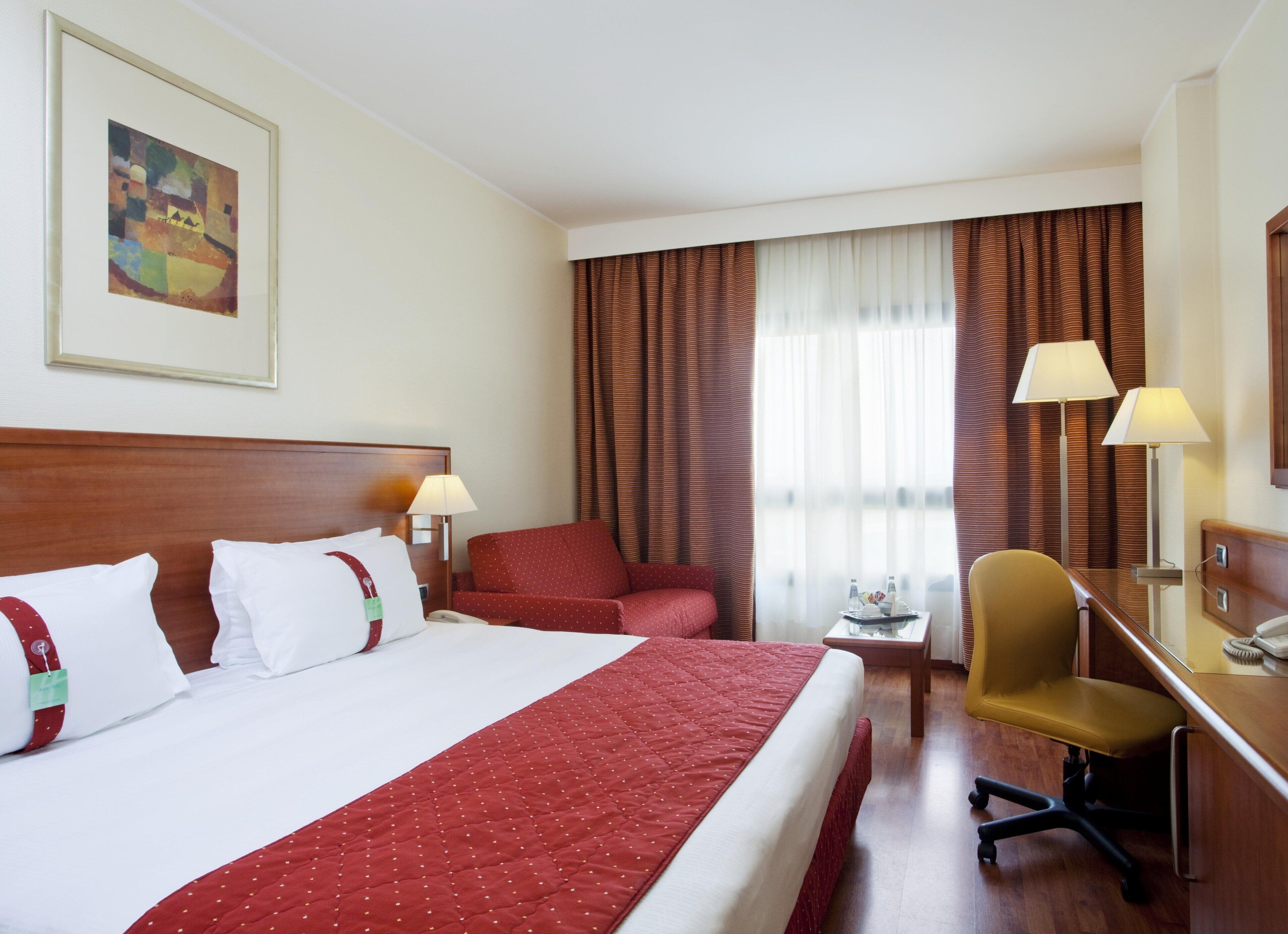 Quarto de hotel com cama, escrivaninha, cadeira, luminária, telefone e um quadro na parede.